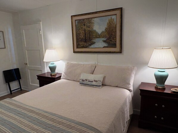 A neatly made bed with a striped blanket, two bedside lamps, a painting of a river, and a door in the background.