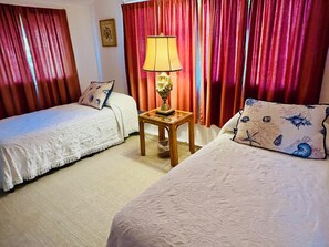 A room with two beds, a nightstand with a lamp, red curtains, and a framed picture on the wall. - Lillie’s Oceanview RUSTIC Boho Surf Cottage 
Steps to the beach & fun! (Summerland)