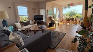 A spacious living room with a sofa, coffee table, and TV. There is a dining area with chairs and a table. The room has large windows and a view of greenery outside. - Lillie’s Oceanview RUSTIC Boho Surf Cottage 
Steps to the beach & fun! (Summerland)