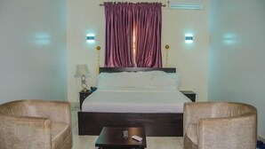A hotel room with a bed, two armchairs, a small table, a nightstand, a lamp, and purple curtains. - Peridot Hotel and Suites (Lagos)