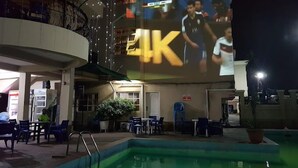 An outdoor pool area with a large screen displaying a sports event, surrounded by seating and potted plants. - Peridot Hotel and Suites (Lagos)