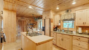 A wooden cabin kitchen with a central island, wooden cabinets, and a window overlooking the forest. - Log Lodge (Woodland Park)