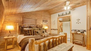 A cozy log cabin bedroom with a wooden bed, two bedside lamps, a rustic bench, and a ceiling fan. - Log Lodge (Woodland Park)