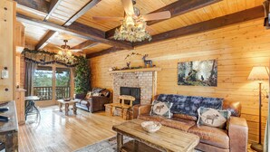 A cozy log cabin interior with a fireplace, leather furniture, and a large window overlooking a deck. - Log Lodge (Woodland Park)