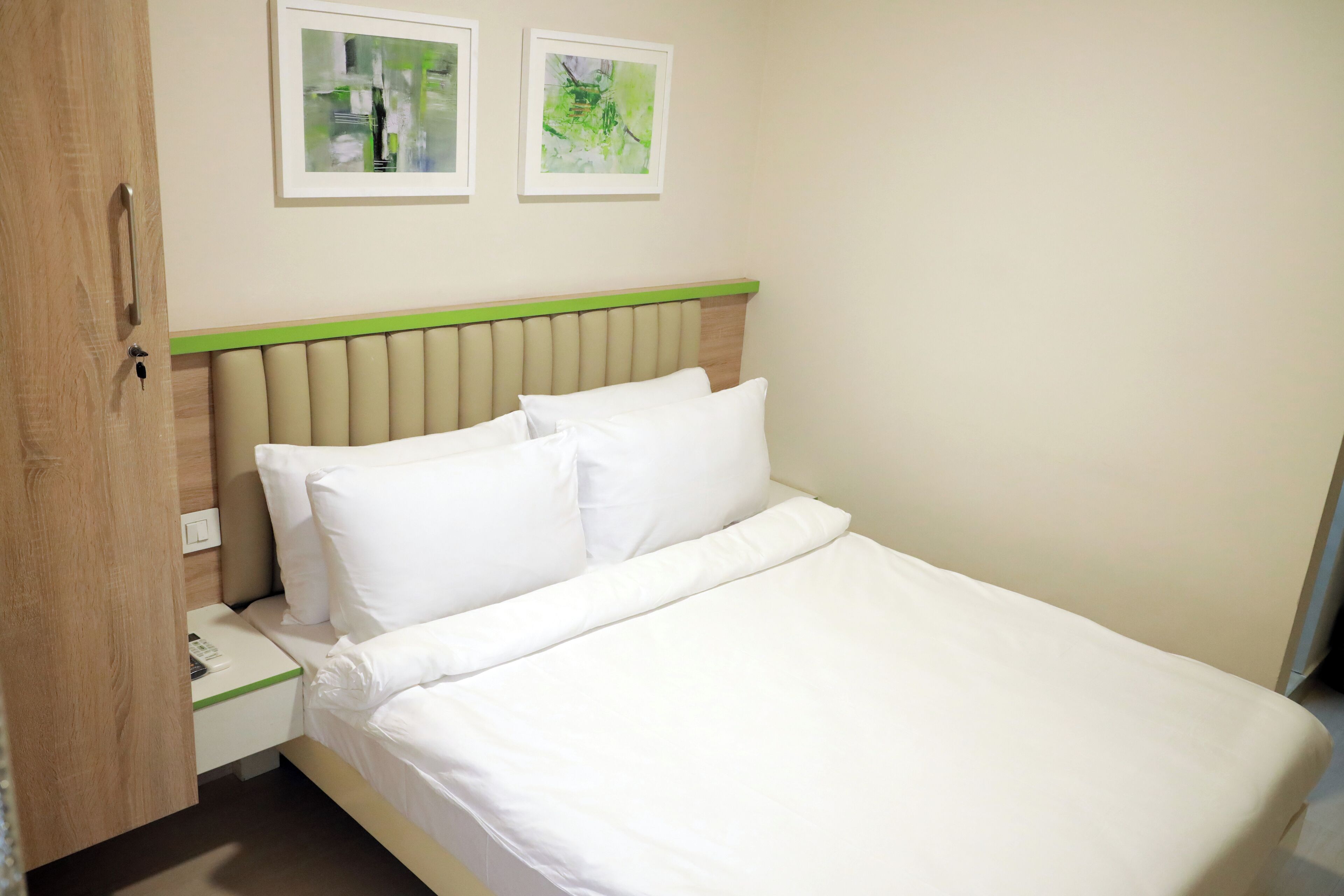 A neatly made bed with white linens, a wooden nightstand with a phone, and two framed abstract paintings on the wall.
