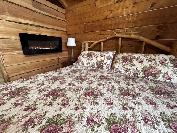 A wooden cabin bedroom with a bed, a wall-mounted fireplace, and a nightstand with a lamp. - Wow! Honeymoon Cabin with Fantastic Views. Very Private and 5 minutes to Parkway (Sevierville)