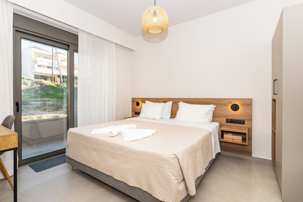 Ein modernes Hotelzimmer mit einem großen Bett, einem Holzkopfteil, einem kleinen Nachttisch, einer Wandleuchte und Blick durch eine Schiebeglastür.