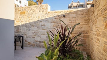 Un muro de piedra con un pequeño jardín, una silla de metal y un edificio al fondo.