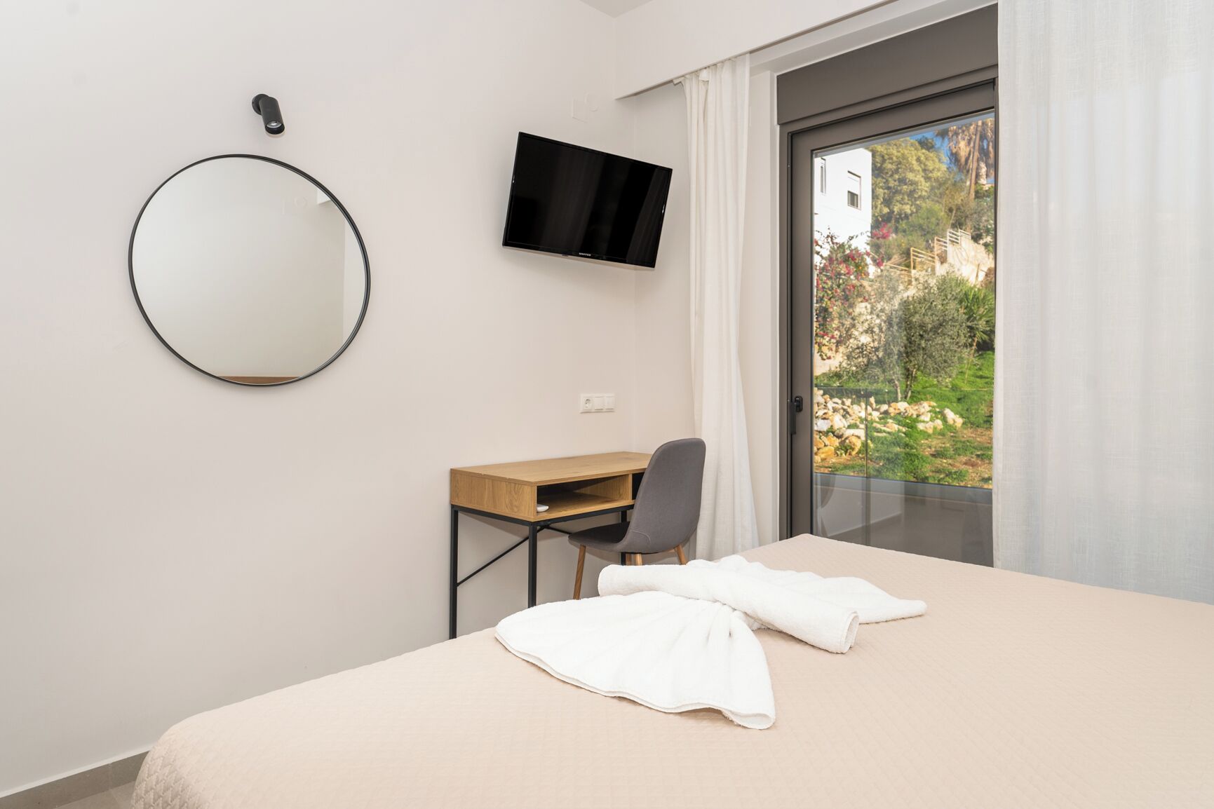 A hotel room with a bed, a desk, a chair, a TV, and a view of greenery outside.