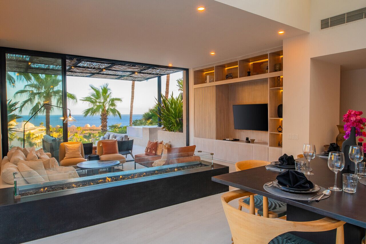 A modern living room with a large glass wall overlooking the sea, furnished with sofas and a dining table set for a meal.