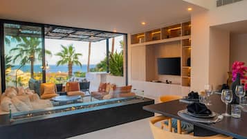 A modern living room with a large glass wall overlooking the sea, furnished with sofas and a dining table set for a meal.