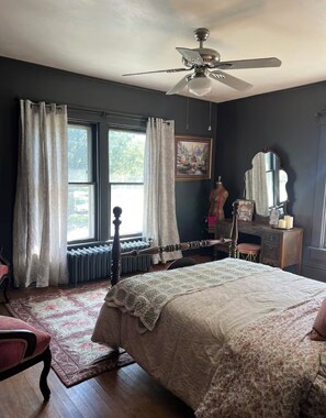 Un dormitorio con una cama grande, una silla de madera, un aparador, un espejo y un ventilador de techo.