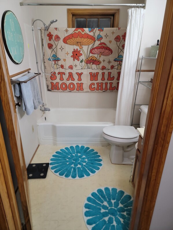 A bathroom with a bathtub, toilet, and a shower. There is a towel rack with a towel hanging on it. The walls have a floral and mushroom pattern with the text "STAY WIL MOON CHIL". There are two blue circular rugs on the floor. - Charming 3-bedroom house with AC, WiFi in pleasant Hardwick.   (Hardwick)