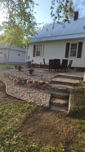 A house with a patio area featuring a stone fire pit, outdoor furniture, and a small garden with potted plants. - Charming 3-bedroom house with AC, WiFi in pleasant Hardwick.   (Hardwick)