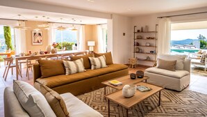 Ein geräumiges Wohnzimmer mit einer Couch, Sesseln, einem Couchtisch und einem Essbereich mit Tisch und Stühlen. Ein Bücherregal und Blick auf einen Pool durch große Fenster.