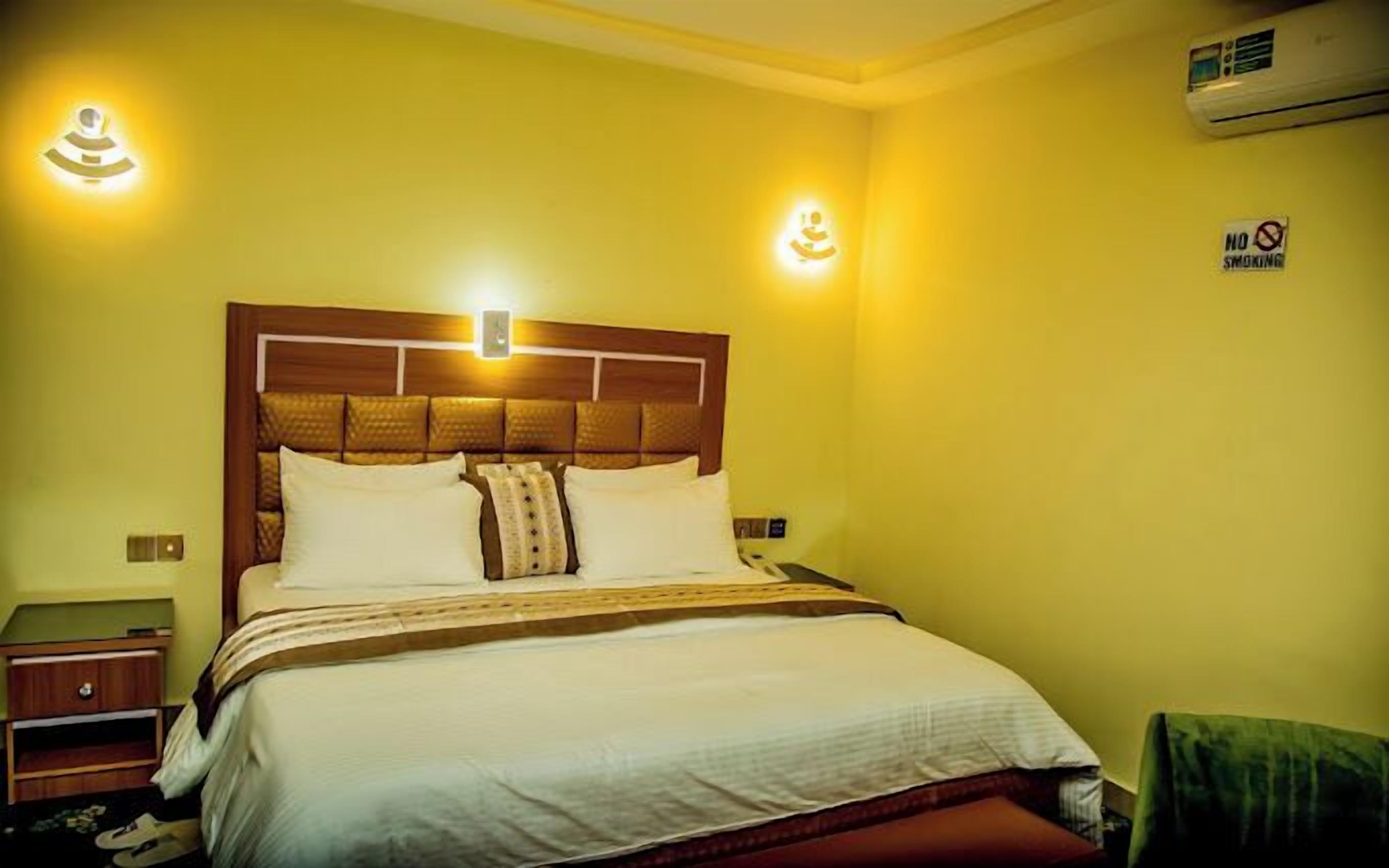 Habitación de hotel con cabecera de madera, cama con ropa blanca, dos lámparas de noche, mesita de noche y un cartel de 'No Fumar' en la pared.