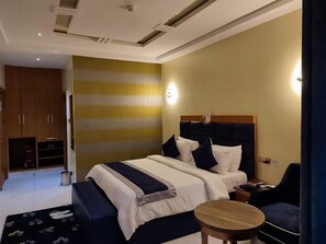 A hotel room with a bed, bedside table, and a small round table. There is a wardrobe and a trash bin in the corner. - Villa Hilton  Hotels and Apartments (Uyo)