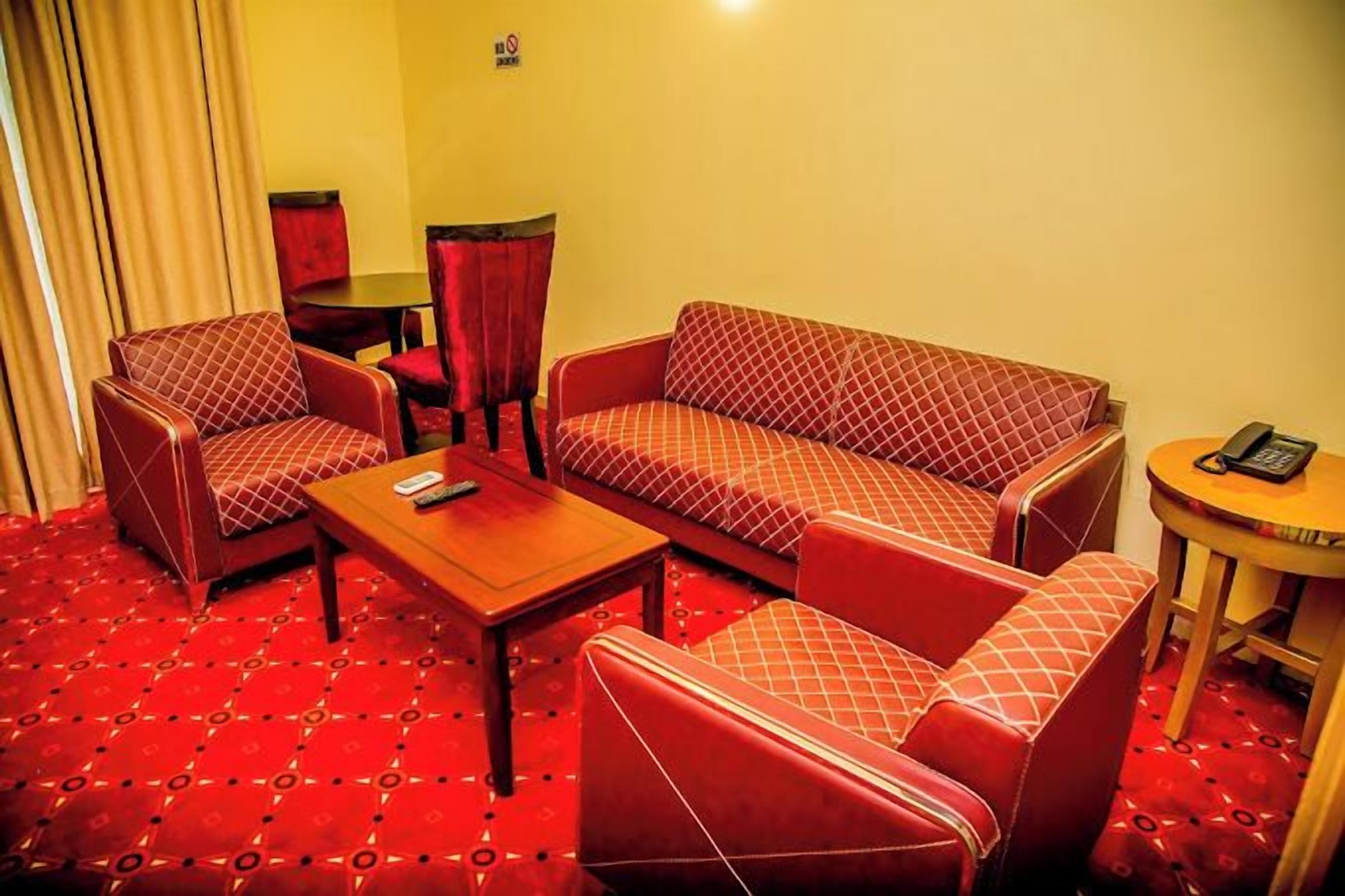 Habitación de hotel con un sofá rojo estampado, dos sillones, una mesita y una mesa redonda con un teléfono.