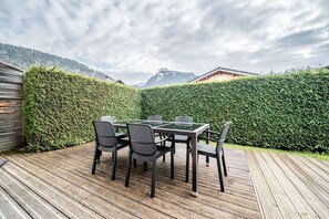 Un terrazzo in legno con tavolo e sedie in vetro, circondato da un alto siepe e con le montagne sullo sfondo.