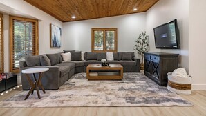A modern living room with a sectional sofa, a coffee table, a TV, and a large window with blinds. - Summit Sanctuary: Urban Elegance mins from Downtown (Flagstaff)