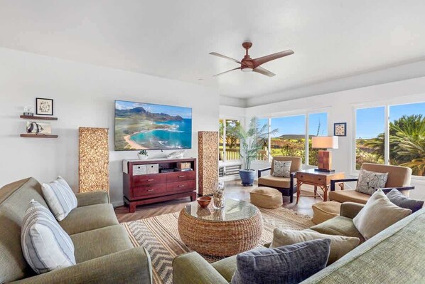A spacious living room with a large window offering a view of the ocean and palm trees. There is a ceiling fan, a television, and a variety of seating including a sofa and armchairs.