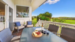 An outdoor patio with wicker furniture, a table with a charcuterie board, wine glasses, and a scenic view of a green landscape. - Whispering Palms – 4BR Home on Kiahuna Golf Course with PBAC Access (Koloa)
