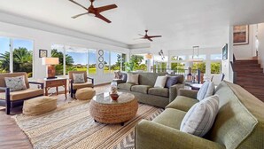 A spacious living room with a large sofa, armchairs, and a coffee table. There are ceiling fans, large windows, and a view of palm trees outside. - Whispering Palms – 4BR Home on Kiahuna Golf Course with PBAC Access (Koloa)