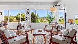 A spacious living room with a large window offering a view of a lush garden. There are four wooden chairs with cushions, a coffee table, and a floor lamp. - Whispering Palms – 4BR Home on Kiahuna Golf Course with PBAC Access (Koloa)