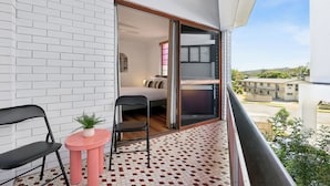 A balcony with a white brick wall, a small pink table with a plant, two black chairs, and a view of a residential area. - Coastal Hideaway on Palm Beach (Palm Beach)