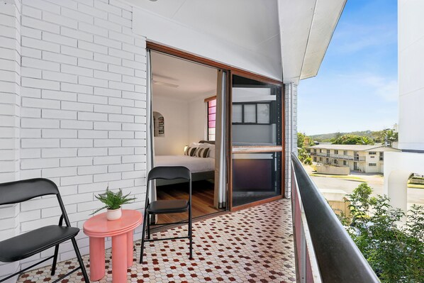 Uma varanda com parede de tijolos brancos, uma pequena mesa rosa com uma planta, duas cadeiras pretas e vista para uma área residencial.