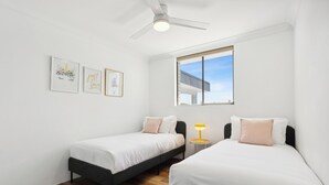 A modern, minimalist bedroom with two single beds, a ceiling fan, and a window with a view of a building. - Coastal Hideaway on Palm Beach (Palm Beach)