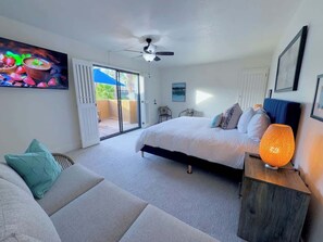 Quarto com cama, sofá, ventilador de teto, mesa de cabeceira com luminária e porta de vidro corrediça para área externa.