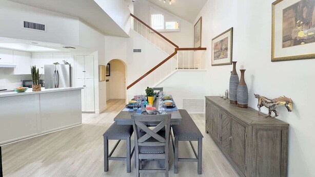 Una cocina moderna con armarios blancos, un comedor con mesa y sillas de madera, y una escalera que sube a un nivel superior.