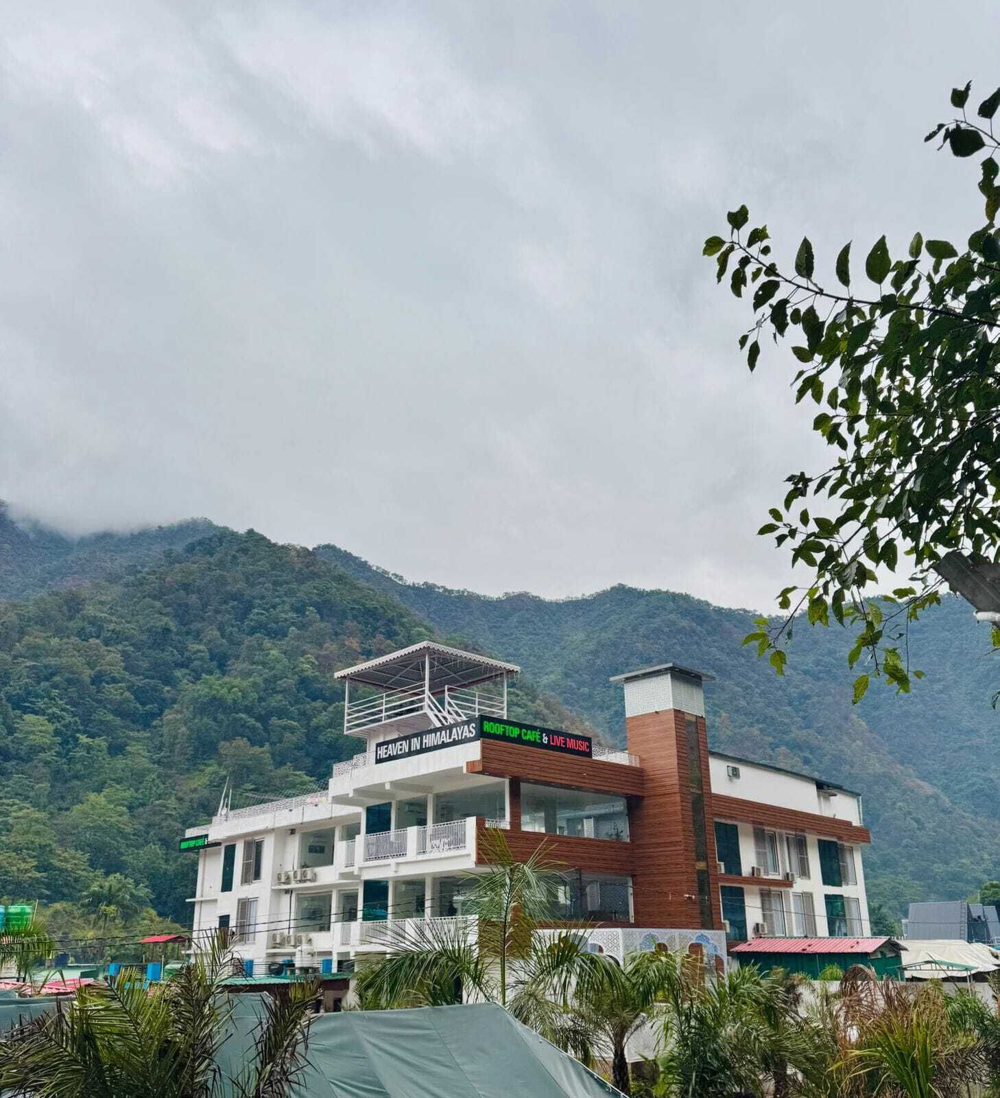 A multi-story building with a rooftop cafe and live music, surrounded by lush greenery and mountains.