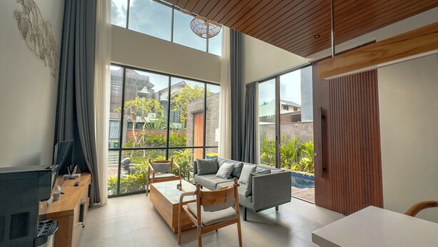 Una sala de estar moderna con sofá, mesa de centro y sillones, ventanas amplias con vistas a un jardín y edificios, y techo de madera.