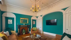 A living room with teal walls, a fireplace, a chandelier, and a television mounted on the wall. - Egerton House Penrith -  a cottage that sleeps 8 guests in 4 bedrooms (Penrith)