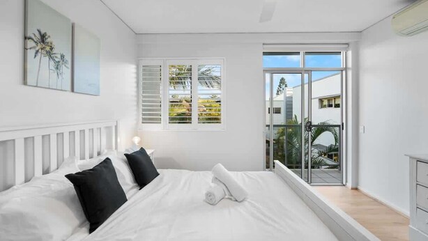 Un dormitorio con cama, paredes blancas, ventilador de techo y vistas a través de un ventanal.