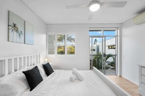 Ein Schlafzimmer mit Bett, weißen Wänden, Deckenventilator und Blick durch eine Glastür.