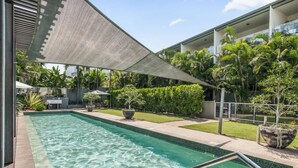 Piscina con zona sombreada, rodeada de follaje y un edificio moderno.