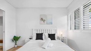 Un dormitorio blanco con cama, una mesita de noche, una planta en maceta y una ventana con persianas.