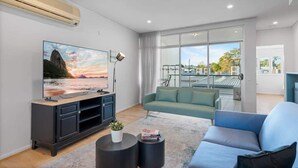 Una sala de estar moderna con un televisor de pantalla plana grande, un sofá, una mesa de centro y vistas al exterior a través de amplios ventanales.
