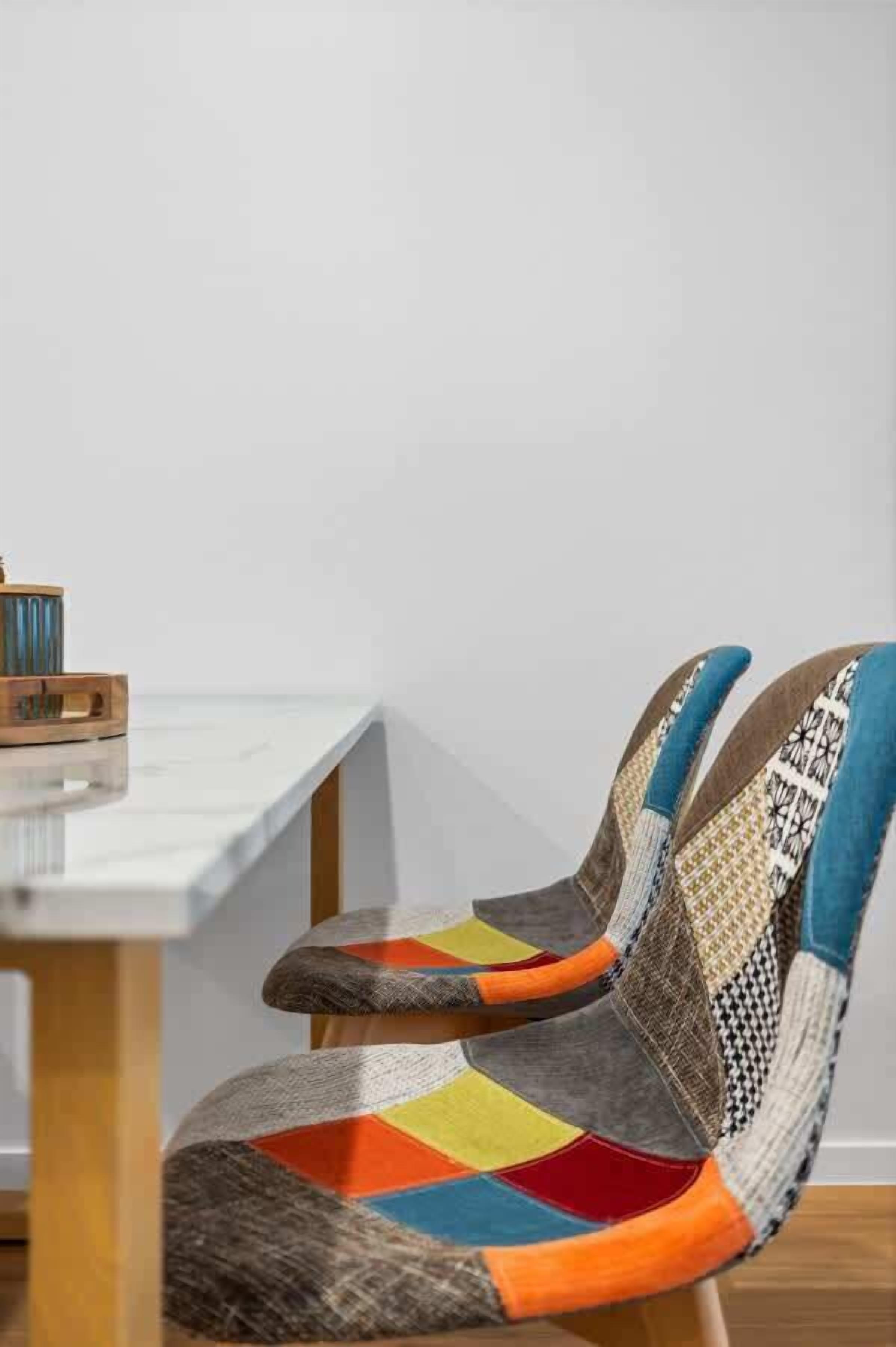 Una silla con un diseño colorido de retazos, una mesita auxiliar de madera con un objeto azul, y una pared blanca.