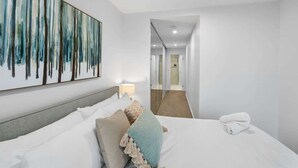 A hotel room with a large bed, a headboard, a wall-mounted abstract painting, a bedside lamp, and a neatly folded towel on the bed.