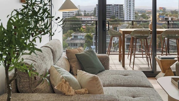 A modern living room with a large sofa, a dining area, and a view of the cityscape through large windows.