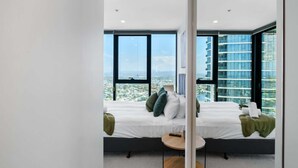 Ein modernes Hotelzimmer mit einem großen Bett, einem kleinen runden Tisch und Blick auf eine Stadtlandschaft durch große Fenster.