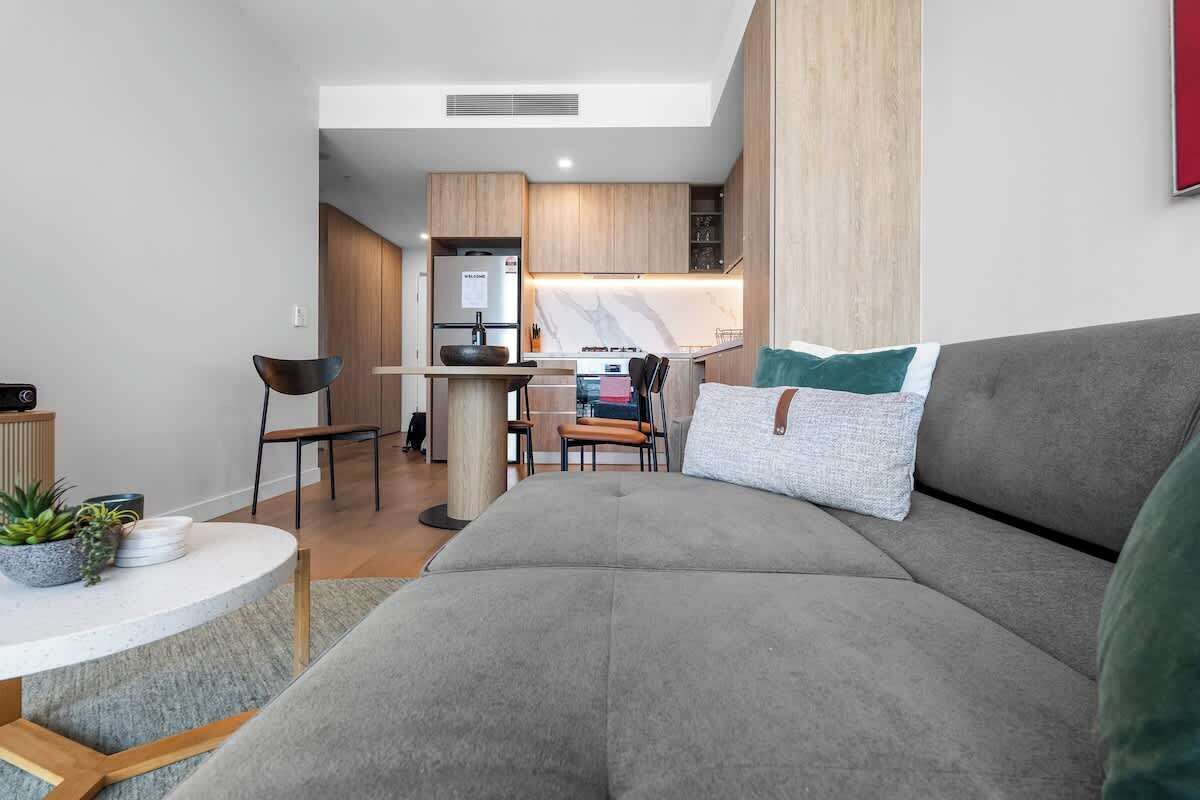 A modern living room with a grey sofa, a small round table with a plant, and a kitchen in the background.