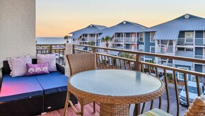 A balcony with wicker furniture, a glass table, and a view of a beachfront hotel.