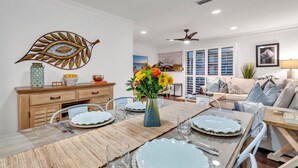 A dining area with a long table set for four, a decorative leaf wall, and a living room with a sofa and a ceiling fan.