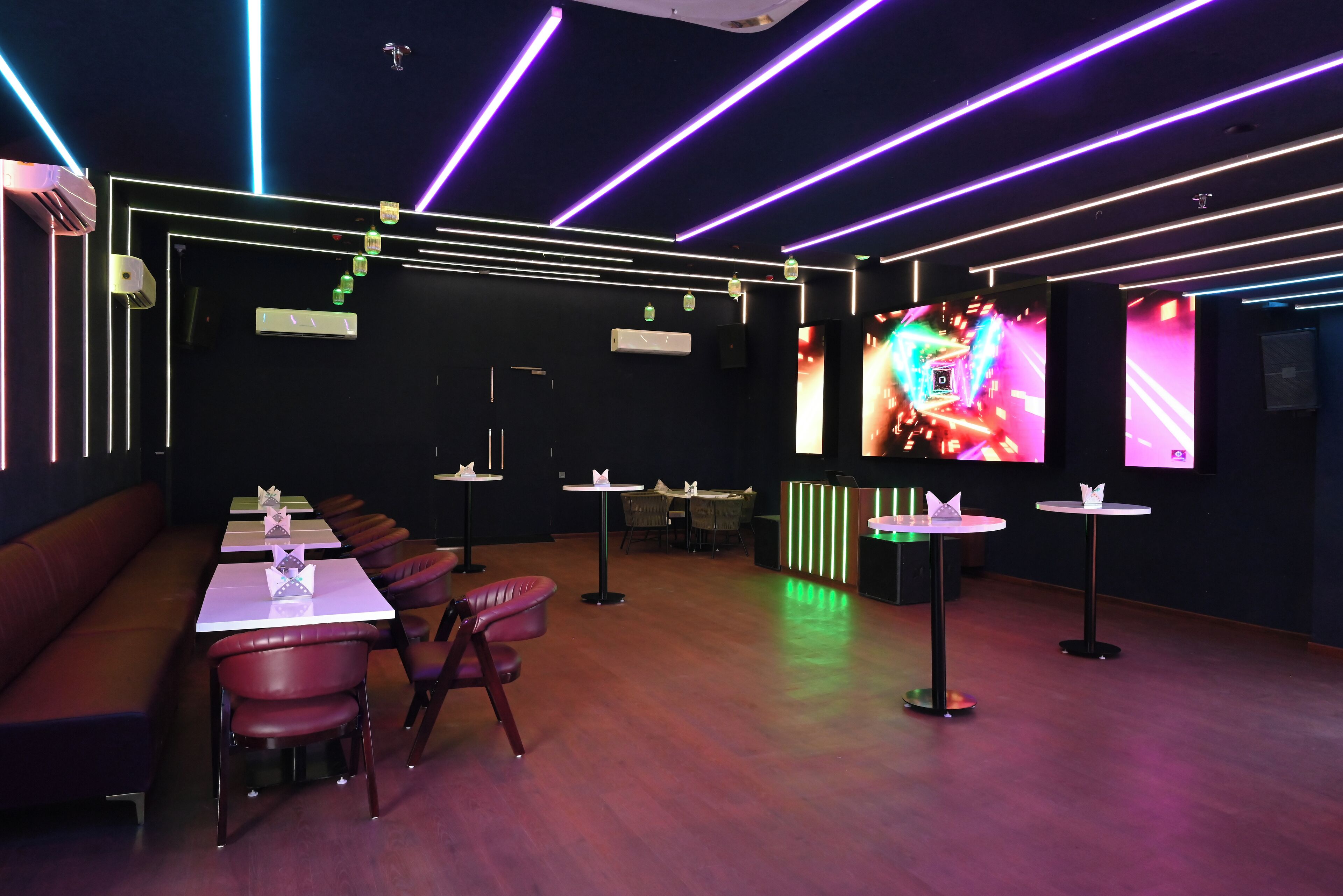 A modern lounge area with a long bench, several tables, and chairs, featuring neon lighting and a large screen displaying colorful visuals.