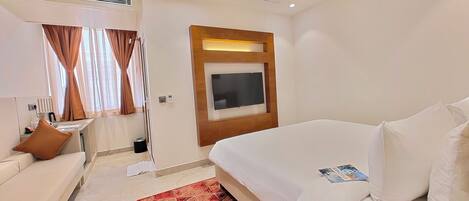 A modern hotel room with a large bed, a sofa, a TV mounted on the wall, and a red patterned rug.